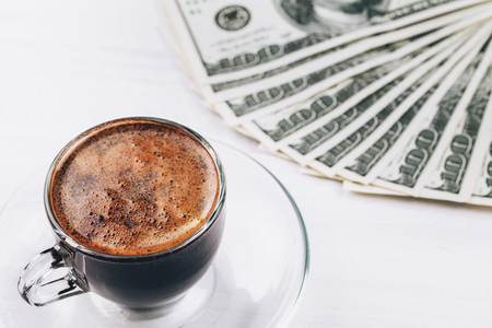 Cup of aromatic espresso in glass with fan of dollars, hundred banknotes, lying on white wooden tableの写真素材