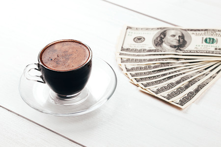 Cup of aromatic espresso in glass with fan of dollars, hundred banknotes, lying on white wooden tableの写真素材