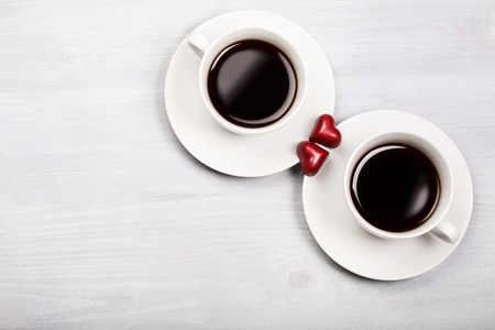 Two white cups of black coffee with two chocolate heart - shaped red sweets. White wooden background. Drinking coffee with love and happy Valentine's day concept.の写真素材