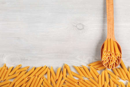 Penne italian pasta lying in wooden spoon and on wooden table, forming textured frame background, closeup.の写真素材