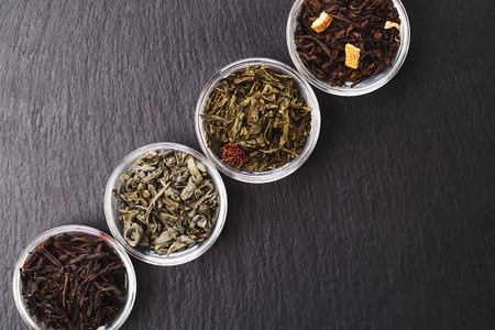 Four bowls with different types of tea: black and green, with fruits are standing on black stone textured backgroung. Place for your text.の写真素材