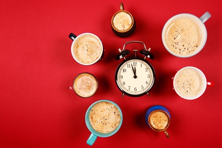 Many different cups of coffee and cappuccino and big alarm clock on red background. Too much caffeine in one dayの写真素材