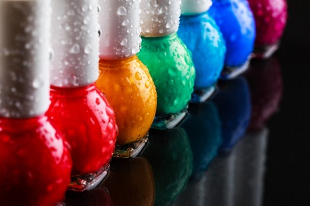 Group of bright nail polishes sorted like a rainbow. Bottles with water drops are standing in a row on black stone background with reflection. Beauty concept.の写真素材