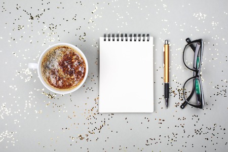 Notebook, coffee cup, smartphone and eye glasses are lying on grey festive background with little...