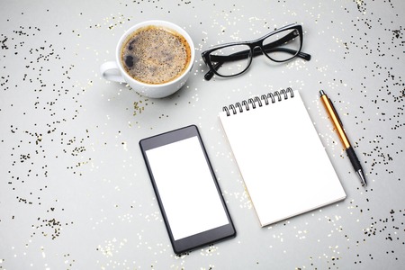 Notebook, coffee cup, smartphone and eye glasses are lying on grey festive background with little golden sparkles. Stuff for business, top view. Place for text.の写真素材