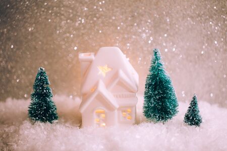 Beautiful christmas card. Little house with windows and light inside and three pines are standing in toy snowy forest on sparkling silver backgroundの写真素材