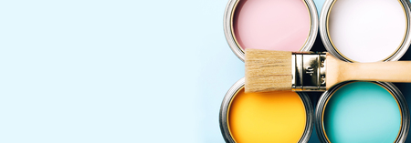 Banner of renovation concept. Brush with wooden handle on opened cans on blue pastel background. Yellow, white, pink, turquoise colors. Macro.の写真素材