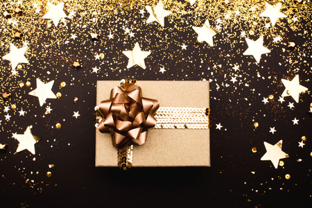 Gift box with golden bow on black background with decoration and sparkles. Flat lay. Festive concept.の写真素材