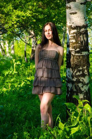 Brunette stands near the birch in forest. girl walking on the green forest. girl in short summer dress in nature .の写真素材