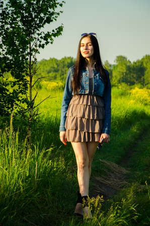 Brunette walks on a green meadow with sunglasses. beautiful brunette girl in a short summer dress walking on green grass path .の写真素材
