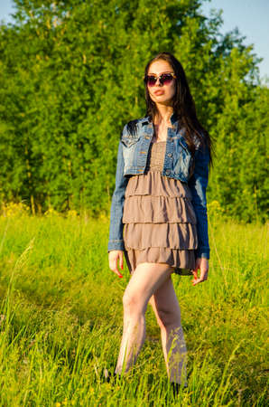 Brunette walks on a green meadow with sunglasses. beautiful brunette girl in a short summer dress walking on green grass path .の写真素材