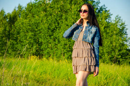 Brunette walks on a green meadow with sunglasses. beautiful brunette girl in a short summer dress walking on green grass path .の写真素材