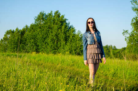 Brunette walks on a green meadow with sunglasses. beautiful brunette girl in a short summer dress walking on green grass path .の写真素材