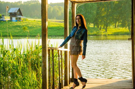 girl standing on a wooden pier . brunette in a short summer dress is standing on tiptoes on the pier at the pond.の写真素材
