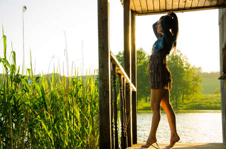 girl standing on a wooden pier . barefoot brunette in a short summer dress is standing on tiptoes on the pier at the pond.の写真素材