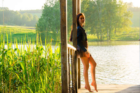 girl standing on a wooden pier . barefoot brunette in a short summer dress is standing on tiptoes on the pier at the pond.の写真素材