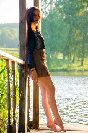 girl standing on a wooden pier . barefoot brunette in a short summer dress . It is standing on tiptoes on the pier at the pond.の写真素材