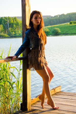 girl standing on a wooden pier . barefoot brunette in a short summer dress is standing on tiptoes on the pier at the pond.の写真素材