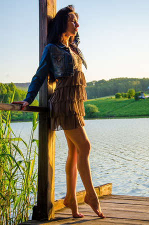girl standing on a wooden pier . barefoot brunette in a short summer dress is standing on tiptoes on the pier at the pond.の写真素材