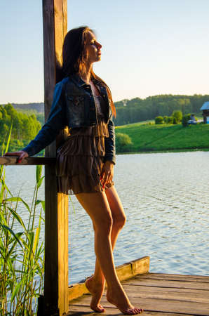 girl standing on a wooden pier . barefoot brunette in a short summer dress is standing on tiptoes on the pier at the pond.の写真素材