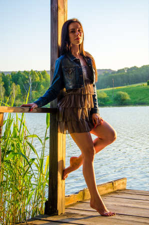 girl standing on a wooden pier . barefoot brunette in a short summer dress is standing on tiptoes on the pier at the pond.の写真素材