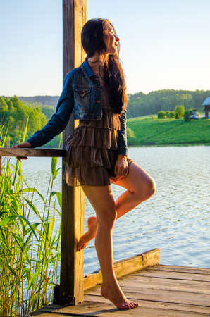 girl standing on a wooden pier . barefoot brunette in a short summer dress is standing on tiptoes on the pier at the pond.の写真素材