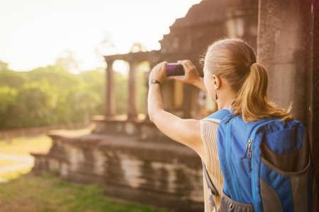Young female tourist with blue backpack and smartphone taking picture of the corner entries of the outside structure of the ancient mysterious temple complex Angkor Wat. Siem Reap, Cambodia.の写真素材