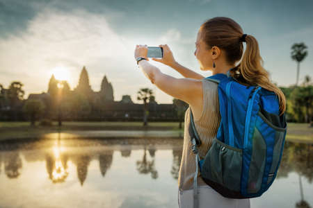 Young female tourist with backpack and smartphone taking picture of the ancient temple complex Angkor Wat reflected in pond at sunrise on background of rising sun. Siem Reap, Cambodia.の写真素材