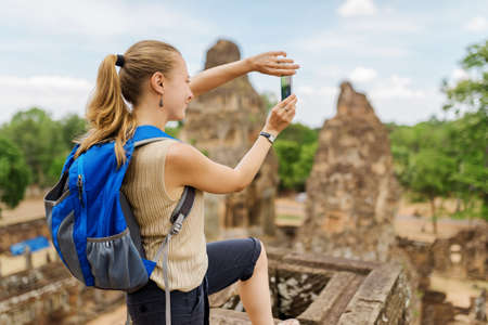 Closeup view of young female tourist with blue backpack taking picture with smartphone from the top of ancient Pre Rup temple Khmer in Angkor. Siem Reap, Cambodia.の写真素材