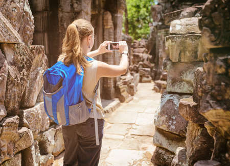 Young female tourist with blue backpack and smartphone taking picture among mysterious ruins of ancient Preah Khan temple in Angkor. Siem Reap, Cambodia. Toned image.の写真素材