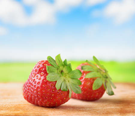 Fresh ripe red juicy strawberries on wooden board. Green field and blue sky with white clouds in background. Healthy eco sweet food rich in vitamins. Popular product of organic farming.の写真素材