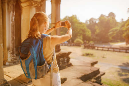Young female tourist with blue backpack and smartphone taking picture among mysterious ruins of ancient temple complex Angkor Wat in morning sun. Siem Reap, Cambodia.の写真素材