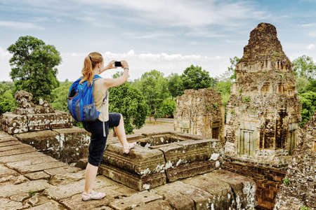 Young female tourist with blue backpack taking picture with smartphone of the gopura from the top of ancient Pre Rup temple Khmer in Angkor on blue sky background. Siem Reap, Cambodia.の写真素材