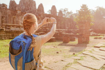 Young female tourist with blue backpack and smartphone taking picture among mysterious ruins of ancient Angkor Thom in Siem Reap, Cambodia. Toned image.の写真素材