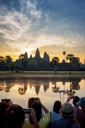 Asian tourists with smartphones taking picture of ancient Angkor Wat at sunrise. Siem Reap, Cambodia. Towers of Angkor Wat reflected in lake at dawn. Angkor Wat is a popular tourist attraction.のeditorial素材