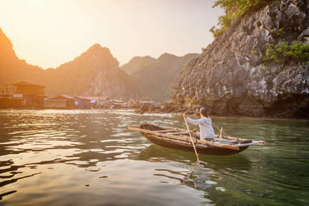 Vietnamese female in traditional boat returns to floating fishing village in the Halong Bay at sunset. The Gulf of Tonkin, the South China Sea, Vietnam. Karst towers-isles are visible in background.の写真素材