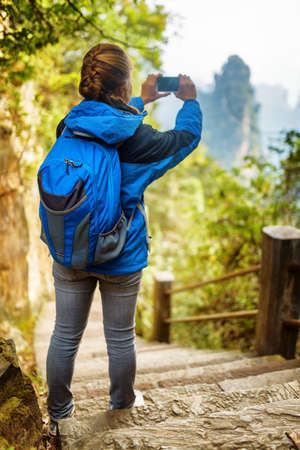 Young female tourist with blue backpack standing on stone stairs among green foliage and taking photo of beautiful mountain view in the Zhangjiajie National Forest Park, Hunan Province, China.の写真素材