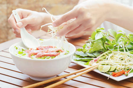 Young woman sprinkles sprouts to the Pho Bo in street cafe of Vietnam. The Pho Bo is a traditional...