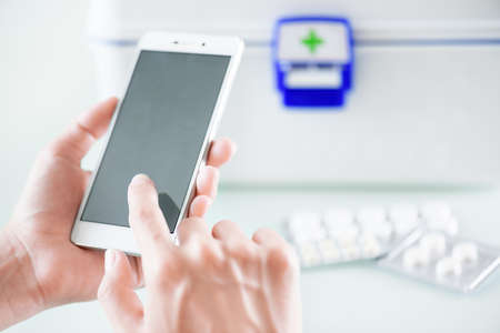 Closeup view of womans hands using smartphone for buying drugs in online pharmacy and getting medical consultation at home. White plastic box with first aid kit and pills in blister packs on table.の写真素材
