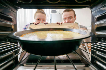 Young woman and young man looking at cheesecake into oven in kitchen. View from inside of the oven. Housewife and her husband holding the oven door.の写真素材