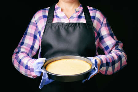 Freshly baked cheesecake in hands of woman on black apron background. Woman wearing checkered shirt. Focus on the cheesecake. Popular sweet dessert.の写真素材