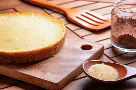 Freshly baked cheesecake on wooden board. Homemade sweet dessert, family meal. Cane sugar in wooden spoon and cocoa powder in glass jar on table.の写真素材