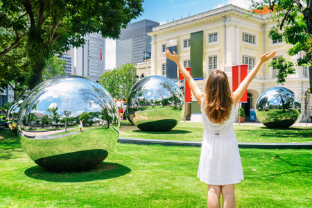 Girl with raised arms to the sky among amazing large mirror balls enjoying the sun and beautiful cityscape in downtown of Singapore. Young woman wearing white dress on green lawn. Freedom concept.のeditorial素材