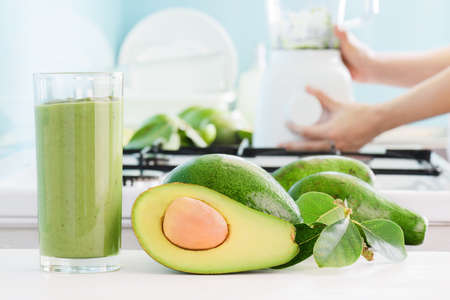 Fresh avocado smoothie and ripe green avocados on kitchen table. Hands of woman using blender are...