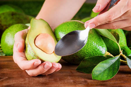Hands of woman holding spoon and half of fresh ripe avocado. Green avocados on wooden table. Healthy...