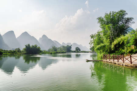Beautiful view of the Yulong River and scenic karst mountains at Yangshuo County of Guilin, China. Amazing green hills reflected in water. Small bamboo raft is visible by a pier at right side.の写真素材