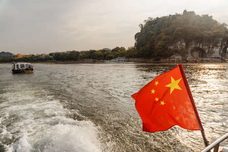 The flag of the Peopleâs Republic of China (the Five-star Red Flag) fluttering on boat sailing on the Li River at downtown of Guilin at sunset. The Elephant Trunk Hill is visible in background.の写真素材