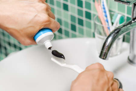 Close-up view of hands of young woman squeezing black charcoal toothpaste on a white toothbrush at bathroom. Colorful toothbrushes are visible in background. Whitening toothpaste. Dental health.の写真素材