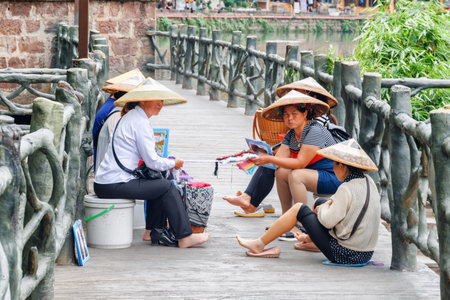 Fenghuang, China - September 22, 2017: Chinese sellers of souvenirs in traditional bamboo hats resting at walkway in Phoenix Ancient Town. Fenghuang is a popular tourist destination of Asia.のeditorial素材