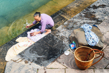 Fenghuang, China - September 22, 2017: Top view of old Chinese woman washing clothes in the Tuojiang River at Phoenix Ancient Town (Fenghuang County). Traditional Chinese wicker backpack.のeditorial素材